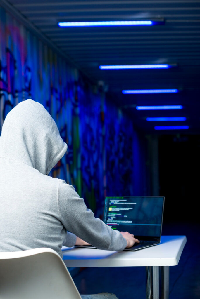 Person in a gray hoodie typing on a laptop with code on the screen, symbolising hacking activity and cybersecurity threats.